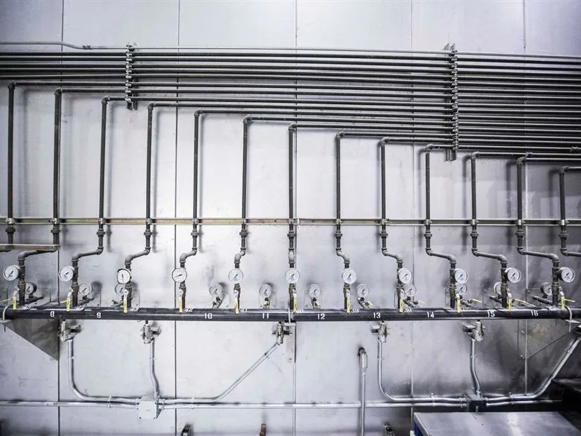 Galeria de tubulação em aço inox com uma sequência de medidores e registros alinhados na parede.